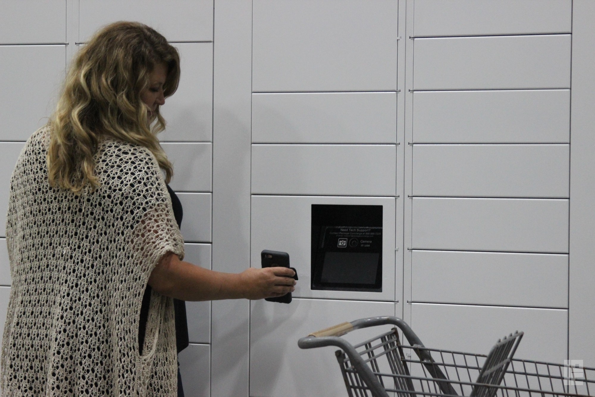 A woman with a white shawl is holding up her smartphone to a white Package Concierge automated locker system to scan her barcode and retrieve her delivered package