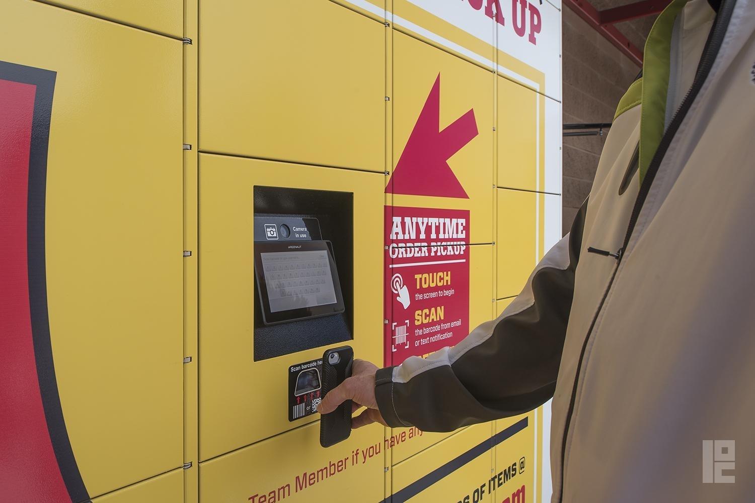 A yellow and red Package Concierge® automated locker system is displayed. A man in a jacket is seen holding up his phone to the barcode scanner to retrieve his package from the locker system